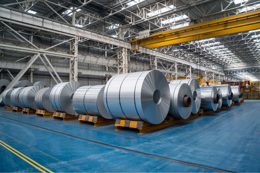 Large industrial warehouse with rows of shiny metal coils on yellow pallets under a high ceiling with metal beams and skylights.