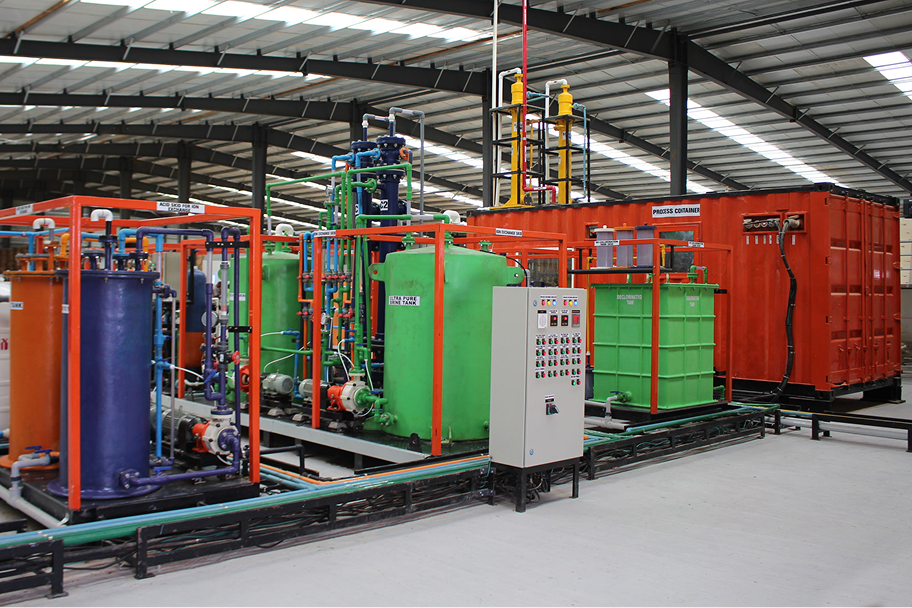 Industrial facility interior with colorful tanks, pipes, a control panel, and red containers under a metal roof.